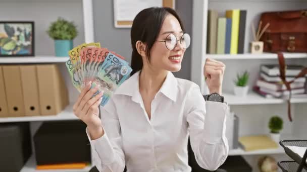若い中国人女性を笑い 自信を持ってオフィスに親指を向け オーストラリアのドル紙幣を保持する喜びの喜びの表現が口を開いた 屋内設定で楽観的で陽気な — ストック動画