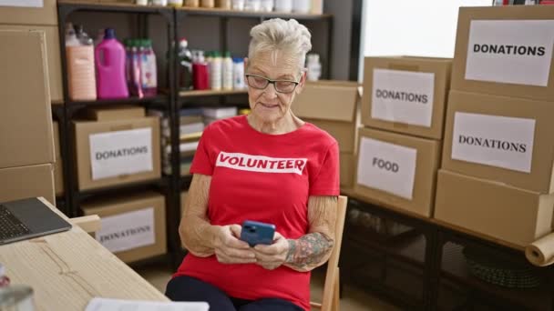 快乐的 头发灰白的老年女性志愿者 一边带着捐款 一边用智能手机在慈善中心发短信 自信地微笑着 — 图库视频影像