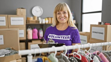 Gülümseyen, güzel, sarışın orta yaşlı bir kadının, hayat dolu bir hayır kurumunda bağışlanan kıyafetleri kontrol etmesi ve not alması çok iç açıcı.