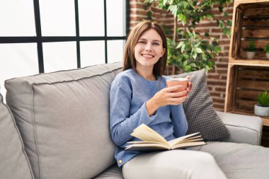 Genç sarışın kadın evde kahve içip kitap okuyor.