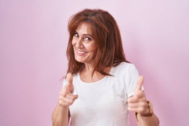 Pembe arka planda duran orta yaşlı kadın mutlu ve komik yüzlü parmaklarını kameraya doğrultuyor. iyi enerji ve titreşimler. 