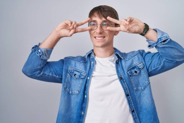 Beyaz, sarışın, gözlüklü, barış sembolü takan, yüzünde parmakları olan, gülümseyen, zaferi gösteren bir adam. 