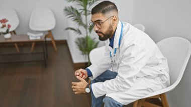 Bearded man in white coat sitting indoors, gripping stress ball tightly in a modern waiting room clipart