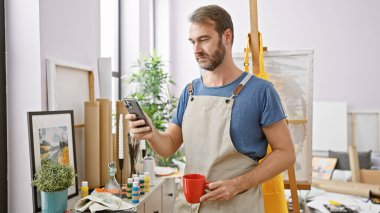 Sanat stüdyosunda telefon ve kahve tutan önlüklü yakışıklı, orta yaşlı bir adam.