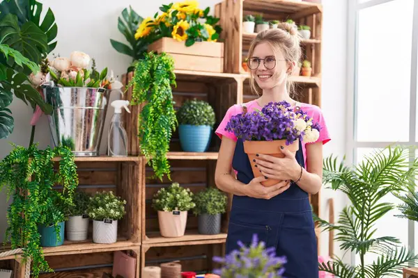年轻的金发碧眼的女花匠微笑着 满怀信心地捧着植物在花店里 — 图库照片
