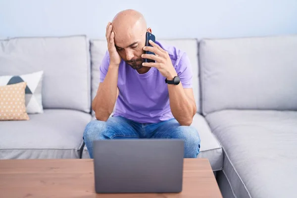 Young bald man talking on smartphone with worried expression at home