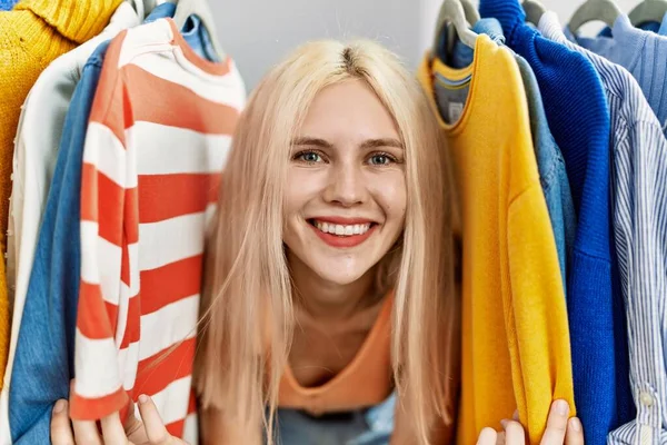Young blonde woman customer smiling confident between clothes at clothing store