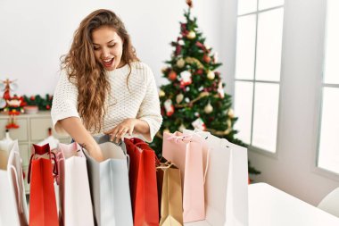 Young beautiful hispanic woman looking shopping bags standing by christmas tree at home clipart