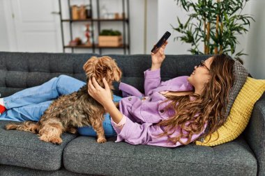 Young beautiful hispanic woman using smartphone lying on sofa with dog at home