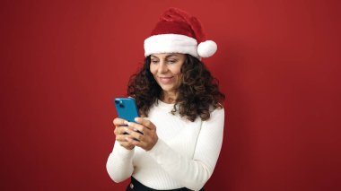 Orta yaşlı İspanyol kadın, izole edilmiş kırmızı arka plan üzerinde akıllı telefon kullanarak Noel şapkası takıyor.