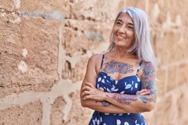 Orta yaşlı, gri saçlı, kolları çapraz duran bir kadın izole taştan arka planda hareketler yapıyor.