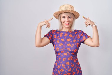 Genç beyaz kadın çiçek desenli elbise ve yaz şapkası takıyor. İki eliyle başını gösteren bir gülümseme. Harika bir fikir ya da düşünce, iyi bir hafıza. 