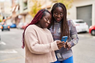 Afro-Amerikalı kadın arkadaşlar, sokakta akıllı telefon kullanarak gülümsüyorlar.