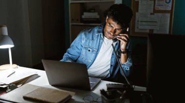 İspanyol adam geç saatlere kadar çalışıyor, bilgisayar ve telefon kullanıyor, odaklanmış bir iş ortamında..