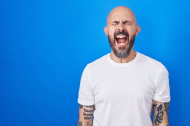 Mavi arka planda dikilen İspanyol dövmeli adam öfkeli ve öfkeli çığlıklar atıyor öfkeyle bağırıyor. öfke ve agresif konsept. 