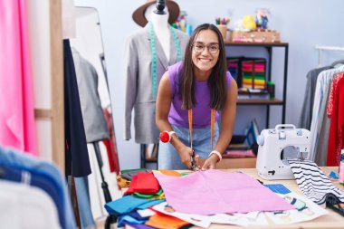 Young beautiful hispanic woman tailor smiling confident cutting cloth at tailor shop