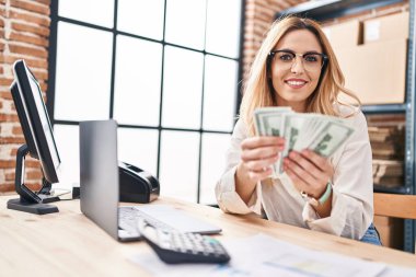 Young blonde woman ecommerce business worker using laptop holding dollars at office