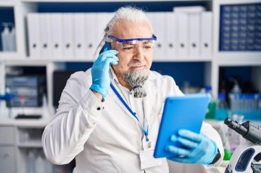 Middle age grey-haired man scientist talking on smartphone using touchpad at laboratory