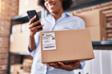 African american woman ecommerce business worker using smartphone holding package at office