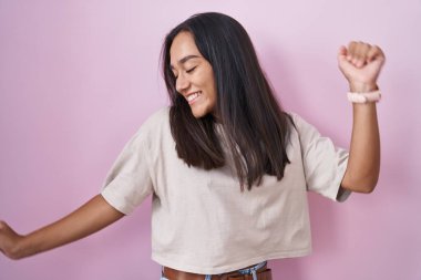 Young hispanic woman standing over pink background dancing happy and cheerful, smiling moving casual and confident listening to music 