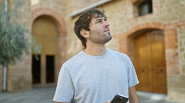 Rahat, sakallı genç bir adam, akıllı telefonuna dalmış, yeşil bir parktaki gökyüzüne bakıyor - mesajlaşmaya dalmış yakışıklı bir yetişkinin portresi, onu dışarıdaki dünyaya bağlayan teknoloji
