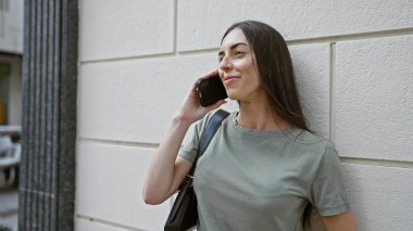 Genç, güzel, İspanyol bir kadın gülümsüyor ve caddede akıllı telefondan konuşuyor.