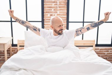 Genç kel adam yatak odasında kollarını esneterek uyanıyor.