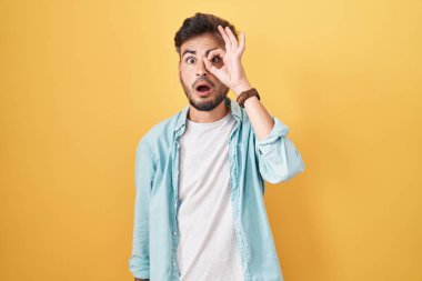Young hispanic man with tattoos standing over yellow background doing ok gesture shocked with surprised face, eye looking through fingers. unbelieving expression. 
