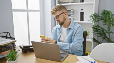 Sakallı ve gözlüklü genç beyaz bir adam modern ofis ortamında akıllı telefonunu kullanıyor..