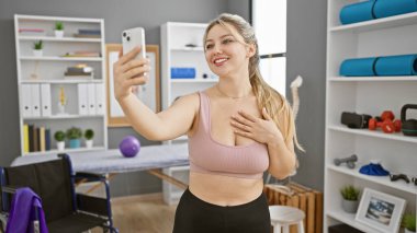 Sarışın kadın fizyoterapi kliniğinde spor malzemeleriyle selfie çekiyor.