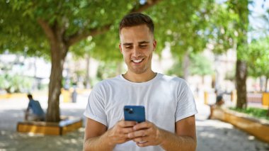 Gülümseyen genç bir İspanyol, yeşil ağaçlı güneşli bir parkta akıllı telefon kullanıyor..
