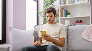 Yakışıklı, sakallı genç bir adam. Kanepede akıllı telefon kullanıyor.