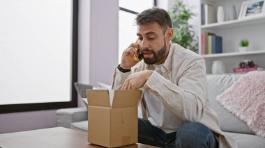 Young hispanic man speaking on the phone unpacking cardboard box at home clipart