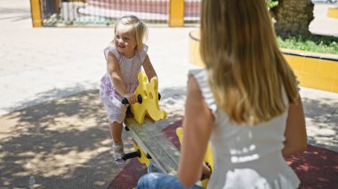 Kafkas anne-kız ikilisi güvenle gülümsüyor, parkta tahterevalliden keyif alıyor.