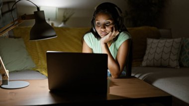 Evdeki kanepede oturan dizüstü bilgisayar ve kulaklık kullanan genç Latin kadın.