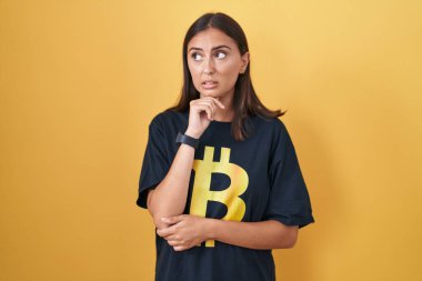 Bitcoin tişört giyen genç İspanyol kadın gergin ve gergin görünüyor. Ağzı tırnak yiyen ellerle. anksiyete. 