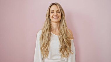 Uzun sarı saçlı, gülümseyen, beyaz bir kazak giyen, pembe arka planda beyaz bir kadın..