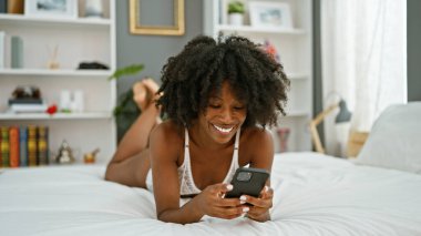 Afro-Amerikan kadın akıllı telefon kullanıyor. Yatakta uzanmış, yatak odasında seksi görünüyor.