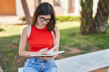 Genç beyaz kadın kitap okuyor, parkta bankta oturuyor.