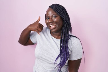 Pembe arka planda duran Afrikalı genç bir kadın gülümsüyor telefonla el ve parmaklarıyla telefon hareketi yapıyor telefonda konuşuyor gibi. iletişim kavramları. 