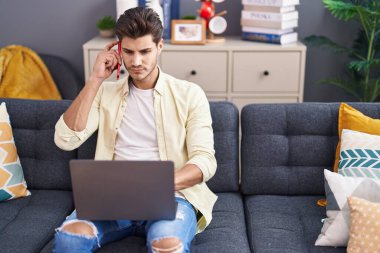Young hispanic man talking on smartphone using laptop at home
