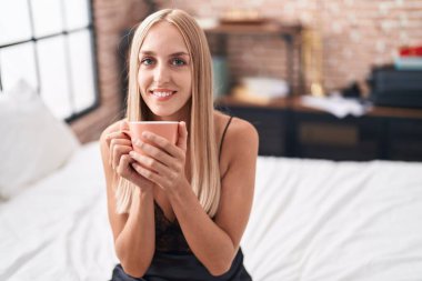 Genç sarışın kadın yatakta bir fincan kahve içiyor.