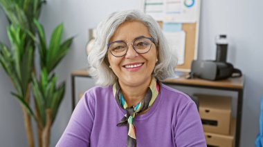 Gri saçlı, gülümseyen olgun bir kadın ofis ortamında gözlük takıyor.