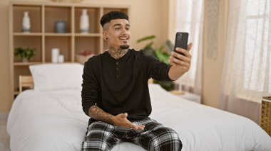 Neşeli genç latin adam evinde yatağında oturmuş video sohbetine daldı, rahat yatak odasında mobil teknolojinin ve dijital bağlantının gücünden yararlandı.