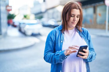 Genç, güzel, iri yarı bir kadın sokakta ciddi bir ifadeyle akıllı telefon kullanıyor.