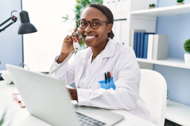 Afro-Amerikalı kadın doktor bilgisayarını kullanarak klinikte telefonla konuşuyor.