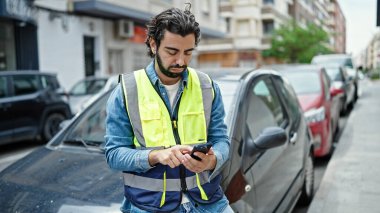 Yansıtıcı yelek giymiş genç bir İspanyol sokakta akıllı telefon kullanıyor.