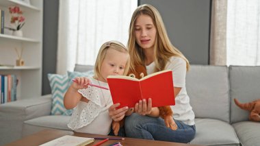Ciddi beyaz anne ve küçük kızı evde oyuncaklarla çevrili bir kanepede oturup kitap okumanın keyfini çıkarıyorlar.