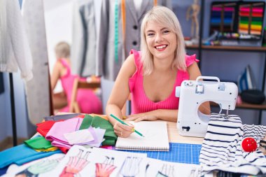 Genç sarışın kadın terzi gülümsüyor oyun odasında defterine çizim yapıyor.