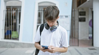 Kulaklıkları ve sırt çantasıyla şehir caddesinde akıllı telefonuyla meşgul olan beyaz bir genç..